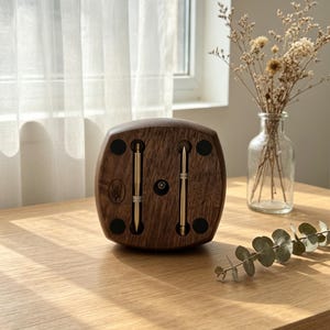 May include: A wooden pen holder with two gold pens inserted. The holder is a dark brown color with a rounded shape and black circular accents. The pens are vertically aligned and centered.