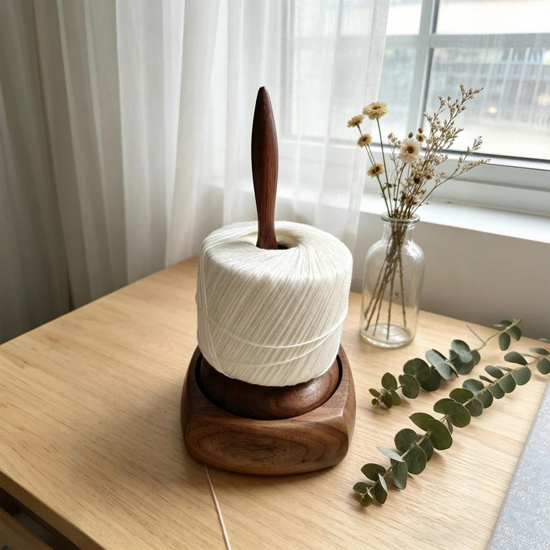 May include: A wooden yarn swift with a dark brown finish, holding a large white yarn ball. The swift has a central wooden spindle and sits on a square base. The yarn is ready for knitting or crocheting.
