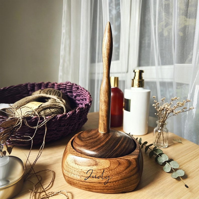 May include: Wooden yarn holder with a dark brown finish. The yarn holder has a round base with a central spindle. The name "Judy" is engraved on the base. A purple basket with yarn is in the background.