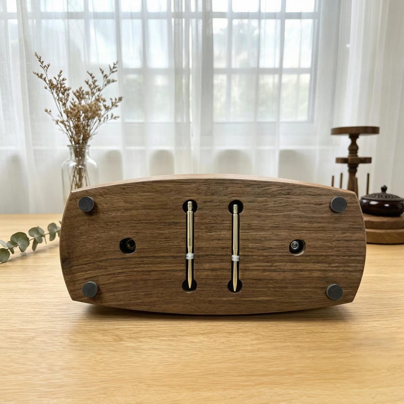 May include: A wooden pen holder with two silver pens. The holder is a dark brown, rectangular shape with rounded edges and small black feet. The pens are inserted vertically into the holder.