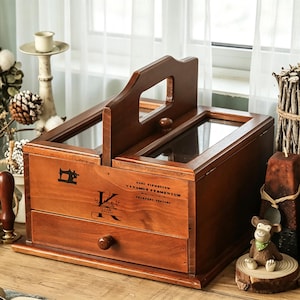 May include: A wooden sewing box with a handle and two glass-topped compartments. The box has a drawer and is decorated with a sewing machine icon and text. The box is a warm brown colour.