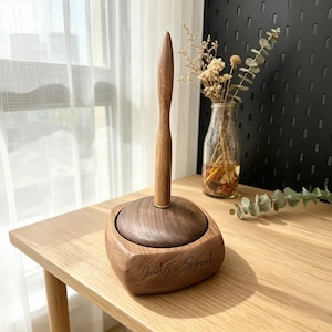 May include: A wooden spool holder with a dark brown base and a lighter brown spindle. The holder is engraved with the text "Judy's Spool". The spool holder is sitting on a light brown wooden table.
