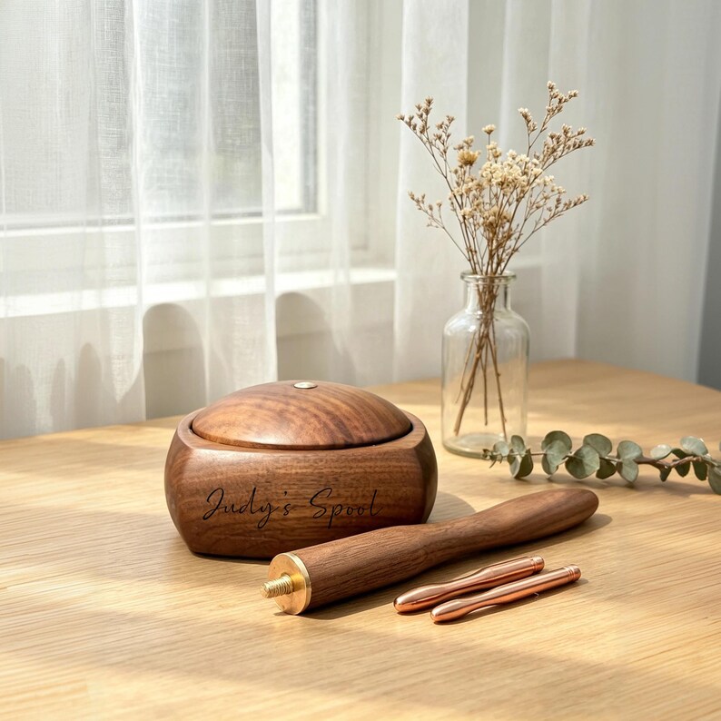 May include: A wooden spool with a lid, engraved with "Judy's Spool", sits on a wooden surface. A wooden tool with a brass end and two smaller copper-colored tools are next to it. A clear glass vase with dried flowers is in the background.