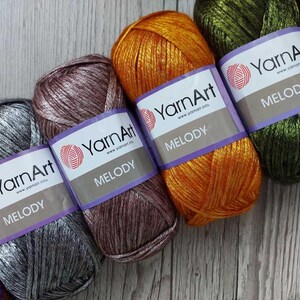 Op de afbeelding: Vier bollen YarnArt Melody garen op een rij, in de kleuren metallic zilver, mauve, feloranje en olijfgroen. Elke bol heeft een wit papieren label met de merknaam YarnArt en de productnaam Melody duidelijk op de voorzijde gedrukt.