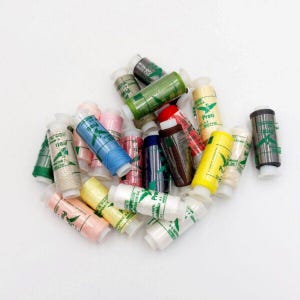 Puede incluir: Una colección de coloridas bobinas de hilo, en tonos de azul, rosa, verde, amarillo y más. Cada bobina está envuelta con hilo y tiene una base de plástico blanco. Las bobinas están dispersas sobre una superficie blanca. El texto "Proti" es visible.