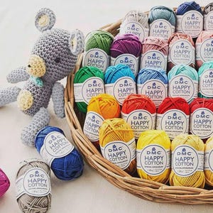 Può includere: Una collezione di gomitoli di filato DMC Happy Cotton colorati in un cestino di vimini, accanto a un orsacchiotto grigio all'uncinetto. I gomitoli di filato sono in vari colori, tra cui verde, giallo, rosso e blu. Le etichette sul filato recitano "DMC Happy Cotton."