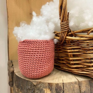 Op de afbeelding: Een kleine, ronde gehaakte mand in oudroze, gevuld met pluizige witte polyester vulling, geplaatst naast een grotere, gevlochten bruine rieten mand met een handvat, alles rustend op een oppervlak van natuurlijk, ruw gesneden hout.