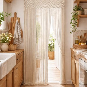 Puede incluir: Una cortina de macramé de color crema cuelga en una puerta, con un diseño geométrico en la parte superior y largos hilos verticales. La cortina complementa los gabinetes y pisos de madera clara de la cocina. Una cesta tejida cuelga en la pared.