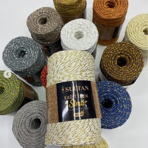 Pode incluir: Uma coleção de doze bobinas redondas de fio Sultan Kağıt İplik Simli em várias cores, incluindo branco, dourado, castanho, cinzento, azul e verde. Cada bobina apresenta um fio metálico entrelaçado e a bobina central tem um rótulo preto.