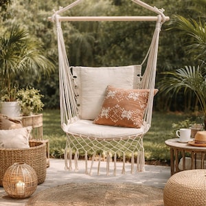 Könnte beinhalten: Ein cremefarbener Makramee-Hängesessel mit Holzstange, verziert mit zwei Kissen. Eines ist cremefarben, das andere rostfarben mit einem weißen Stickmuster. Der Stuhl hängt in einem Garten mit anderer Außendekoration.