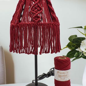 May include: A burgundy macrame lampshade with a black metal base. The lampshade has a fringed bottom and a woven design. A spool of burgundy macrame cord sits next to the lamp. The cord label reads "handiBRAND MACRAME CORD".