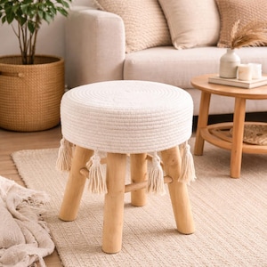 Op de afbeelding: Een ronde, witte kruk bedekt met touw, met houten poten en decoratieve kwastjes. De kruk heeft een neutrale kleur en staat op een beige tapijt. De kruk heeft een diameter van ongeveer 40 cm en is 46 cm hoog.