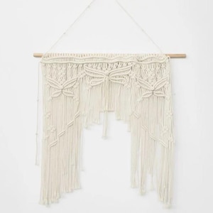 May include: Cream-colored macrame wall hanging. The handcrafted piece features intricate knotting patterns, including floral designs and cascading fringe. It is suspended from a natural wooden dowel, adding a touch of bohemian decor.