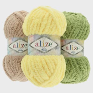 Puede incluir: Tres ovillos de lana Alize Baby Softy Mega en beige, amarillo y verde. Cada ovillo está envuelto con una etiqueta con el nombre de la marca y los detalles del producto. La lana parece suave y esponjosa, ideal para mantas de bebé y otros proyectos.