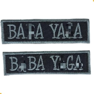 May include: Two rectangular patches with the text "BABA YAGA" embroidered in light gray thread on a black background. The patches have a gray border and are likely for clothing or accessories.