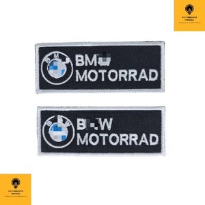 Peut inclure: Deux écussons rectangulaires noirs avec des bordures brodées blanches. Chaque écusson présente le logo BMW dans un cercle blanc avec des accents bleus et blancs, et le mot "MOTORRAD" en texte blanc. Le patch supérieur se lit "BM MOTORRAD", et le patch inférieur se lit "B-W MOTORRAD".