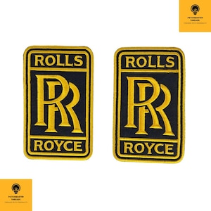Könnte beinhalten: Zwei gestickte Rolls-Royce-Aufnäher. Jeder Aufnäher hat einen schwarzen Hintergrund mit goldfarbenen Buchstaben und einem goldenen Rand. Der Text "ROLLS" steht oben, "ROYCE" unten und ein goldenes "RR" in der Mitte.