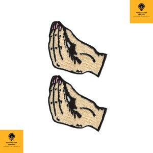 May include: Two embroidered patches featuring a hand gesture with a light skin tone, pink fingernails, and black accents. The patches are outlined in black and have a tan background. The patches are on a white background.