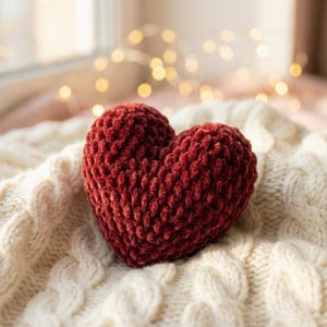 Könnte beinhalten: Ein herzförmiges Dekorationsobjekt in tiefroter Farbe. Das Herz besteht aus einem weichen, strukturierten Material und liegt auf einer cremefarbenen Zopfstrickdecke. Der Hintergrund zeigt weiche, unscharfe Lichter, die eine warme, gemütliche Atmosphäre schaffen.