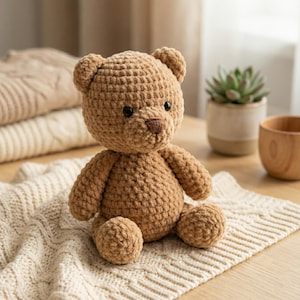 May include: A handmade, light brown crochet teddy bear with black eyes and a brown nose. The bear is sitting on a cream-colored, textured blanket. The bear is a soft, plush toy.