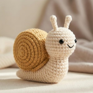 May include: A crocheted snail toy with a tan shell and cream-coloured body. The snail has black eyes and a smiling mouth. The snail's shell is a spiral design. The toy is made of soft yarn.