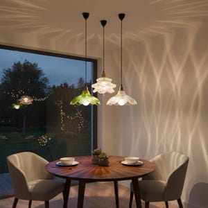 May include: Three pendant lights with textured glass shades hang above a wooden dining table. One shade is green, and the others are clear. The room has a large window and two chairs. The lights cast a pattern on the wall.