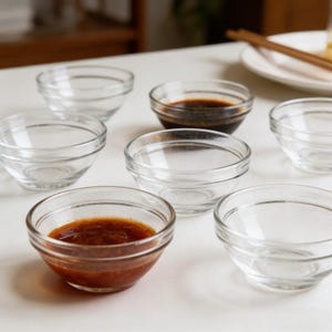 May include: A collection of small, clear glass bowls, some filled with dark brown and reddish-brown sauces. The bowls are arranged on a white surface, suggesting a dining setting. The image highlights the bowls' transparency and the contrast of the sauces.