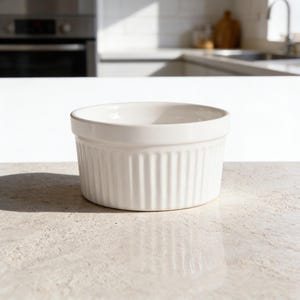 Puede incluir: Un ramequín de cerámica blanca con detalles acanalados verticales. El pequeño plato redondo está diseñado para porciones individuales y es adecuado para hornear o servir. El ramequín está sobre una superficie clara.