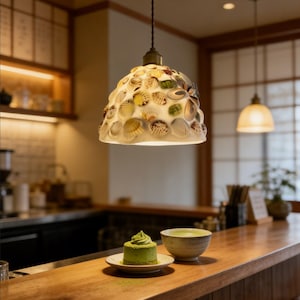 May include: A pendant light fixture with a lampshade adorned with seashells hangs above a wooden counter. A small green cake and a bowl of matcha tea sit on the counter. The light fixture has a bronze-colored top.