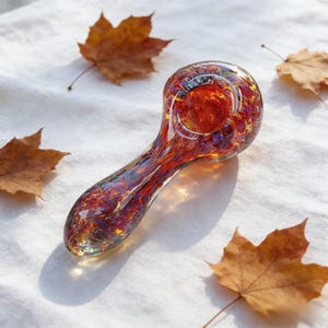 May include: A hand-blown glass pipe with a vibrant blend of red, orange, and yellow hues. The pipe features a rounded bowl and a long, curved stem. Scattered around the pipe are several dried brown maple leaves, creating a natural, autumnal aesthetic.