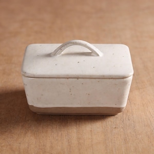 May include: A rectangular ceramic butter dish with a lid and handle. The dish is off-white with speckled detailing and a tan base. The lid has a curved handle for easy lifting. The butter dish is designed for kitchen use.