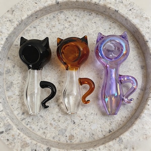 May include: Three glass cat-shaped pipes in black, amber, and iridescent purple. Each pipe features a cat head, a curved tail, and a clear glass body. The pipes are displayed on a textured, light gray surface.
