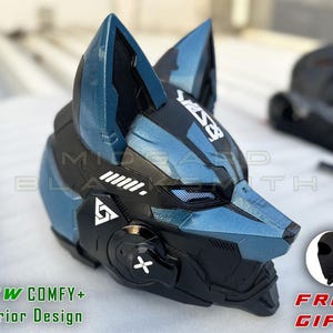 Puede incluir: Un casco con forma de lobo en azul metálico y negro que presenta paneles angulares afilados, orejas puntiagudas y un logotipo geométrico blanco en el lateral. El diseño incluye una zona ocular de malla y un acento lateral circular con una X.