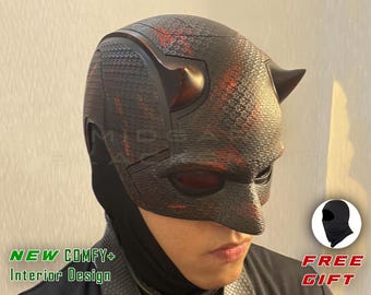 Op de afbeelding: Een zwart en rood getextureerd masker met hoorns, dat lijkt op een superhelden kostuum. Het masker heeft rode oogbedekkingen en wordt gedragen met een zwarte bivakmuts. De tekst "NEW COMFY+ Interior Design" is zichtbaar. Een gratis cadeau van een zwarte bivakmuts is inbegrepen.
