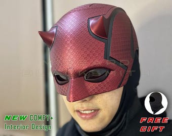 Op de afbeelding: Een rood, getextureerd superheldenmasker met zwarte accenten en twee hoorns. Het masker heeft oogopeningen en wordt over een zwarte bivakmuts gedragen. Tekst op de afbeelding luidt "NEW COMFY+ Interior Design" en "FREE GIFT".