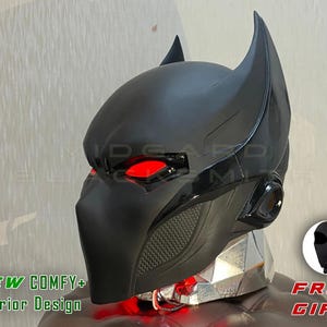 May include: A black helmet with red eye lenses and ear-like points. The helmet features a detailed design with a mesh vent and glossy black trim. The image includes the text "NEW COMFY+ Interior Design" and "FREE GIFT".