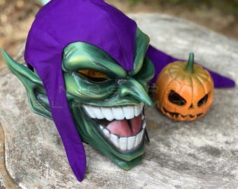 Op de afbeelding: Een groen goblin-masker met een brede, tandige grijns, puntige oren en een paarse stoffen kap. Het masker heeft een glanzende afwerking en staat naast een kleine, oranje, uitgesneden pompoendecoratie. De tekst Midgard Blacksmith is zichtbaar.