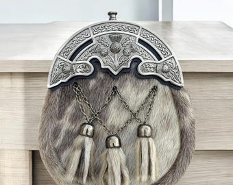 Puede incluir: Un sporran de kilt escocés tradicional con un cantle plateado y un cuerpo de piel. El cantle presenta intrincados nudos celtas y emblemas de cardos. Tres cadenas plateadas con borlas cuelgan del cuerpo del sporran.