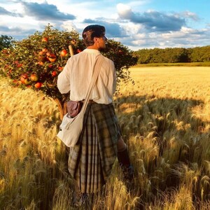 May include: A white long-sleeved button-down shirt, a pleated plaid kilt in shades of green and brown, a brown leather pouch, and a cream-coloured canvas crossbody bag worn over the shoulder, standing in a field of tall golden wheat.