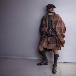 Puede incluir: Una persona vestida con atuendo escocés tradicional, que incluye una falda escocesa a cuadros marrón, una capa a juego y una boina oscura. El atuendo presenta un cinturón de cuero con hebilla dorada, una pequeña bolsa y botas de cuero marrón.