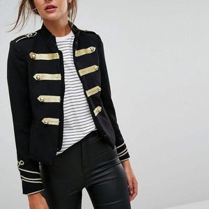 Könnte beinhalten: Schwarze Jacke im Militärstil mit goldenen Akzenten. Die Jacke hat goldfarbene Verzierungen an den Ärmelenden und Schultern sowie goldfarbene Knöpfe. Sie wird über einem gestreiften Oberteil und einer schwarzen Hose getragen.