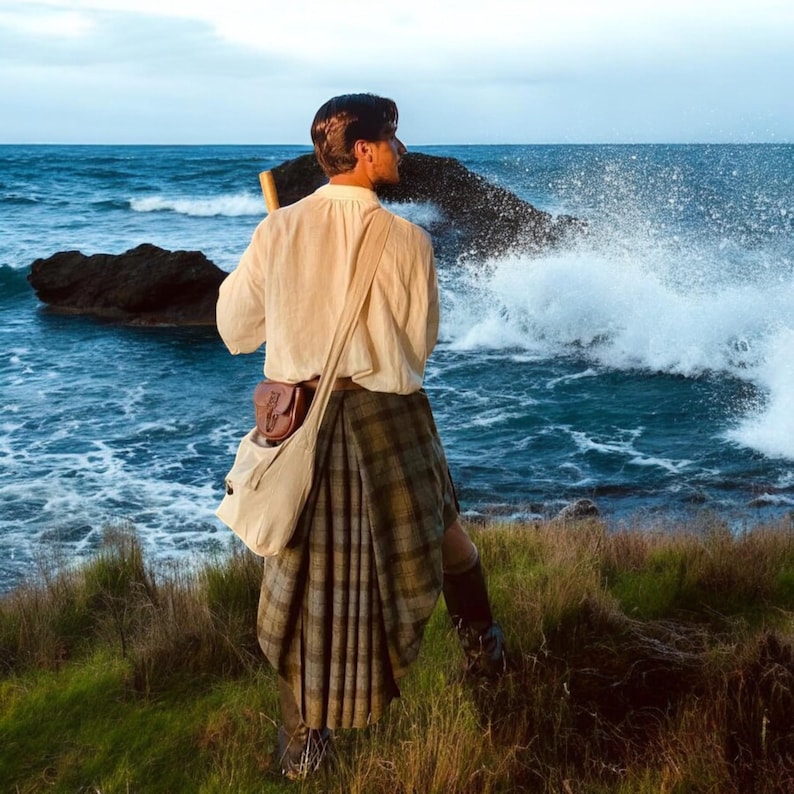 May include: A person wearing a long-sleeved cream-coloured shirt and a pleated kilt with a brown, green, and grey plaid pattern. A light-coloured canvas shoulder bag with a small brown leather pouch attached is worn across the torso over the shirt.
