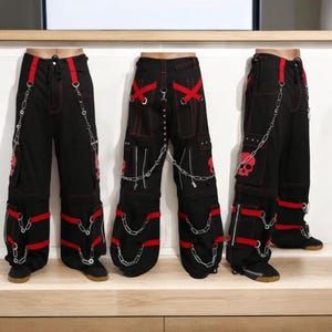 Op de afbeelding: Zwarte cargobroek met rode accenten, met schedelpatches, kettingen en rode stiksels. De broek heeft meerdere zakken en bandjes, met een ontwerp met wijde pijpen. De algehele stijl is edgy en alternatief.
