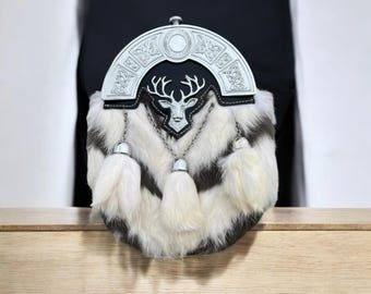 Op de afbeelding: Een traditionele Schotse sporran met een zilverkleurige cantle en een zwarte leren flap. De sporran heeft een hertenkopembleem en witte vacht met zwarte strepen. Drie witte kwasten hangen aan de onderkant.