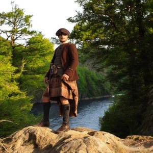 Op de afbeelding: Een persoon draagt een bruine tweed jas, een bruin vest en een traditionele Schotse kilt in een bruin en tan ruitpatroon. De outfit omvat een bijpassende pet, donkere sokken en bruine leren laarzen. De achtergrond toont een rivier en weelderige groene bomen.