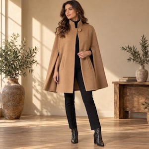 May include: A camel-colored wool-blend cape coat featuring a high collar, a single large button closure at the neck, and arm slits. The garment is worn over a black long-sleeved top and black trousers, paired with black leather ankle boots.