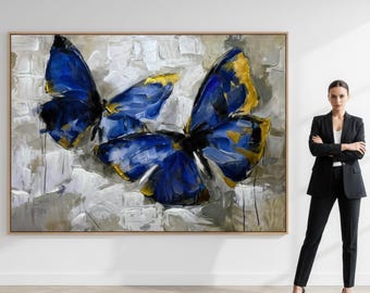 May include: Large framed canvas art featuring three blue and gold butterflies against a textured white and gray background. The artwork is displayed on a wall, showcasing a modern abstract painting style.