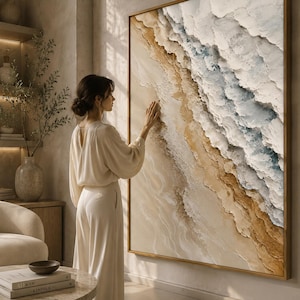 May include: A large square wall art piece featuring a textured abstract landscape with thick impasto layers in shades of sandy beige, cream, and soft blue, framed in a thin gold border, depicting a coastal scene with waves meeting the shore.