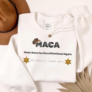 May include: White sweatshirt with the text "MACA" and "Make America Constitutional Again". The design includes a cowboy hat graphic and two gold sheriff badge graphics. A brown cowboy hat is visible in the background.