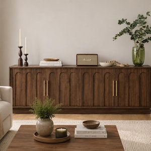 May include: A long, dark wood sideboard featuring vertical arched paneling and slim gold vertical handles. The top surface holds two dark candlesticks, a stack of books, a rectangular speaker, a small bowl, and a green plant in a glass vase.